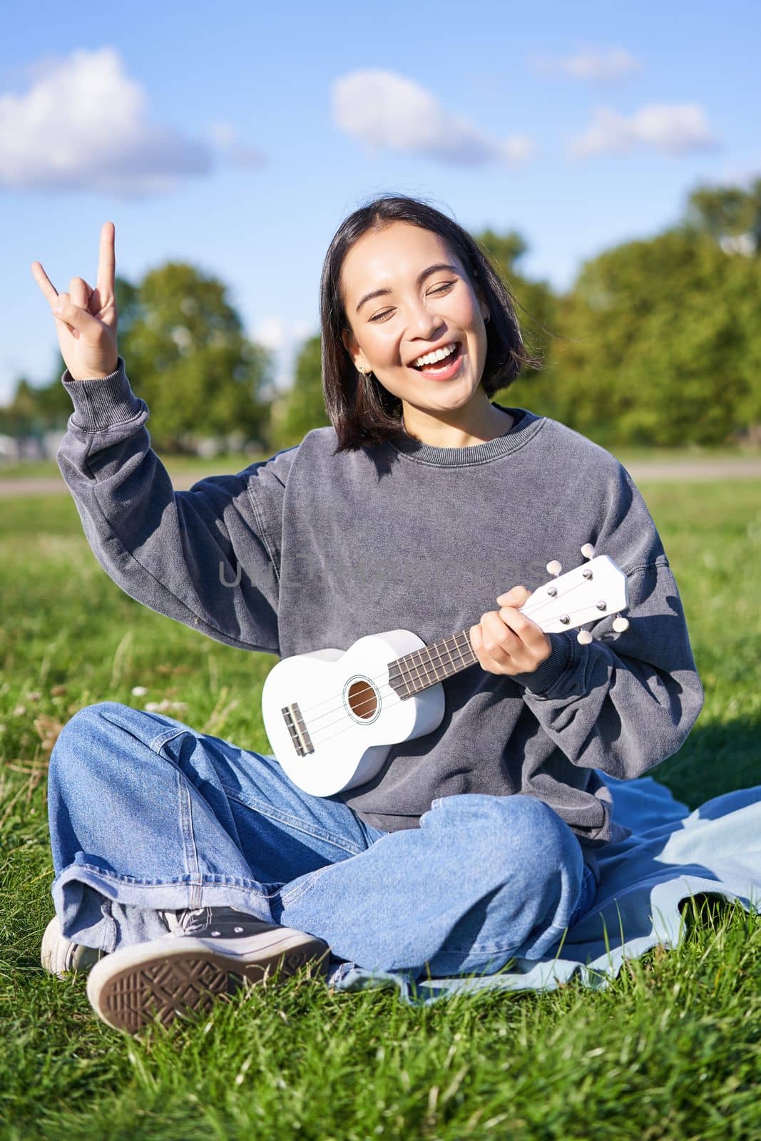 Happy asian girl playing ukulele in park, showing rock n roll, heavy metal horns sign and smiling, having fun outdoors by Benzoix