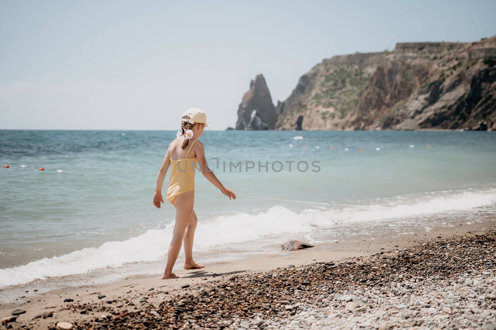 Cute little girl running along the seashore against a clear blue sea and rejoices in the rays of the summer sun. Beautiful girl in yellow swimsuit running and having fun on tropical beach