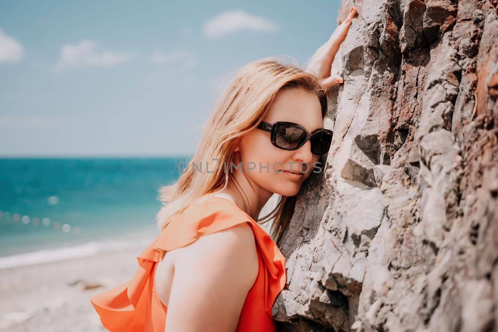 Young woman in red bikini on Beach. Blonde in sunglasses on pebble beach enjoying sun. Happy lady in one piece red swimsuit relaxing and sunbathing by turquoise sea ocean on hot summer day. Close up by panophotograph
