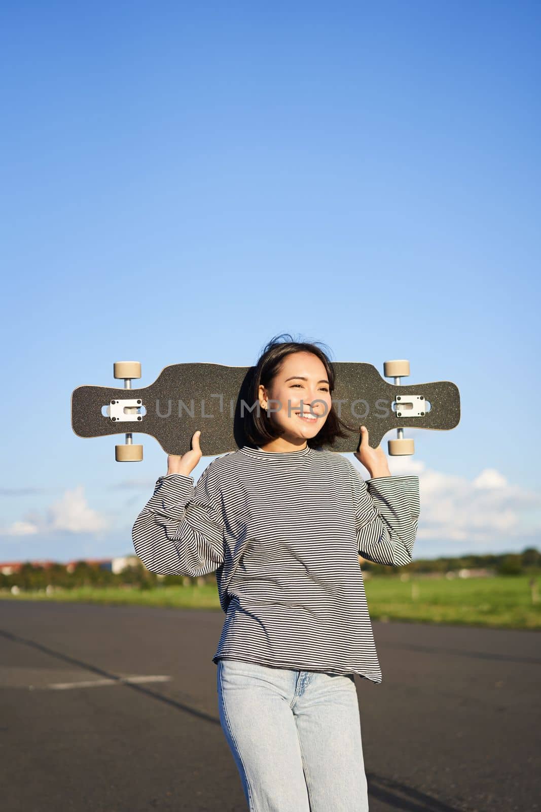 Vertical shot of carefree asian girl with longboard. Young woman skater holding cruiser on her shoulders and walking on road, skateboarding.