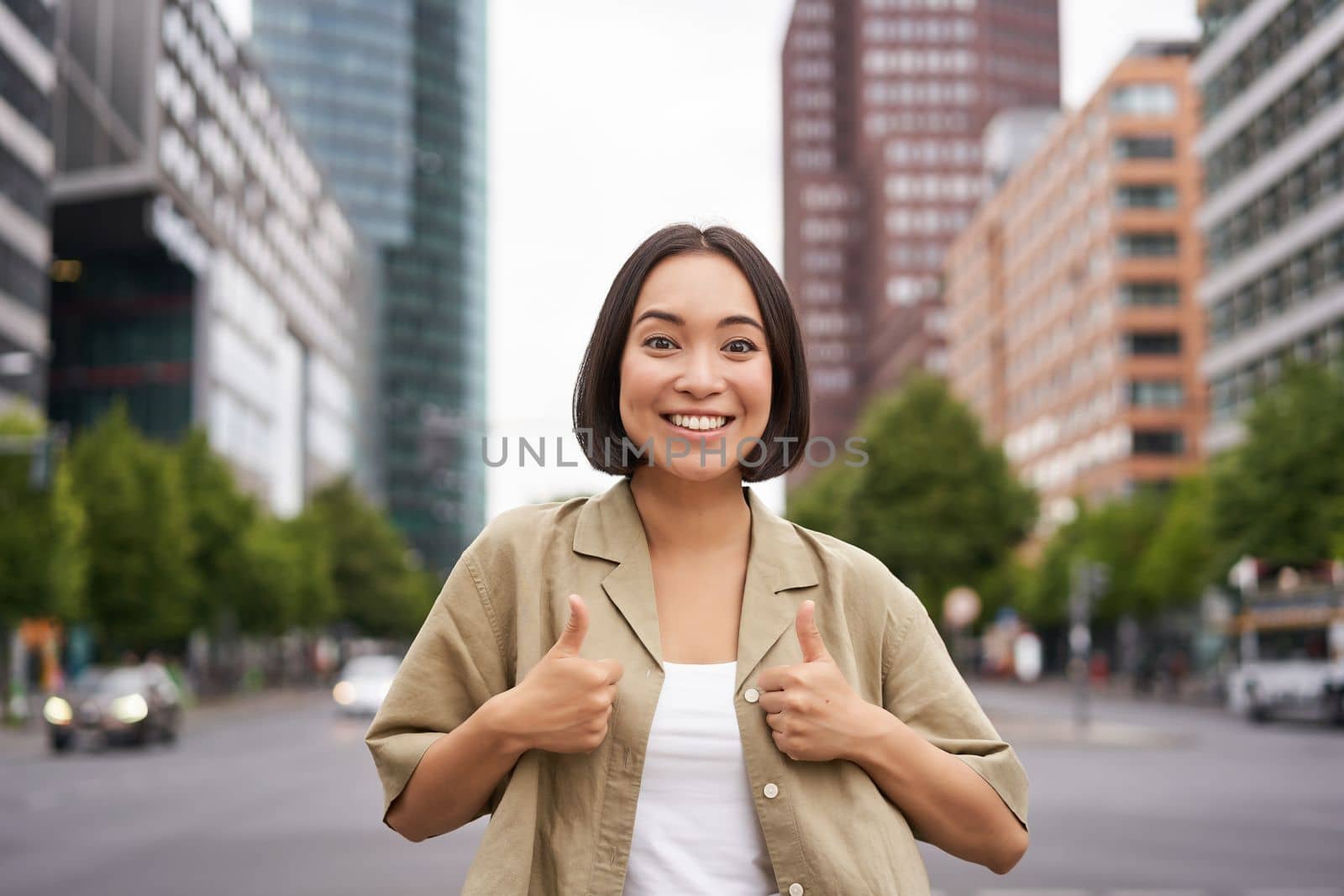 Enthusiastic city girl, shows thumbs up in approval, looking upbeat, say yes, approves and agrees, stands on street.