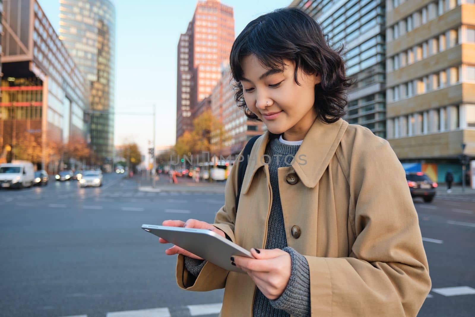 Woman walking on street using digital tablet, holding gadget in hands and looking away, standing in city centre.