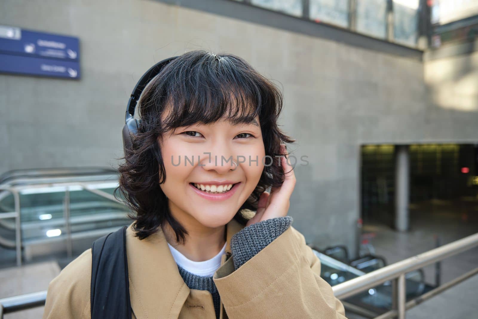 Close up of smiling brunette girl in headphones, listens music, travels around city, commutes to work, stands on a street.