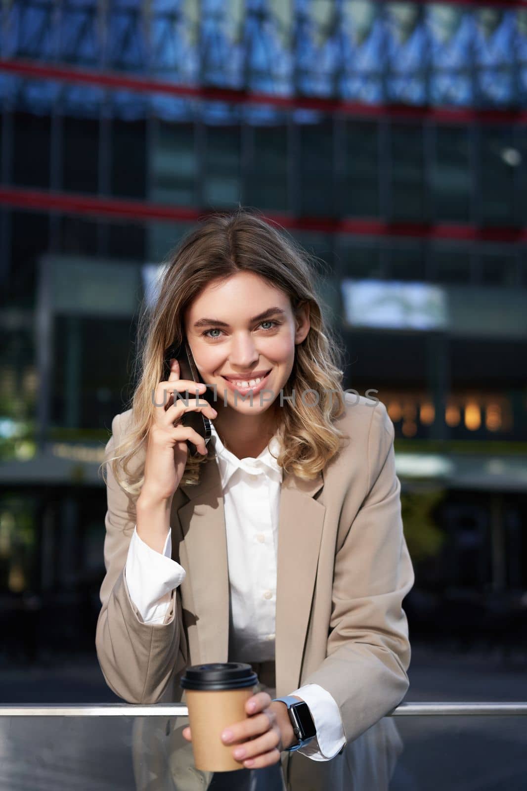 Vertical shot of smiling businesswoman answer mobile phone call, talking on smartphone and drinking coffee.