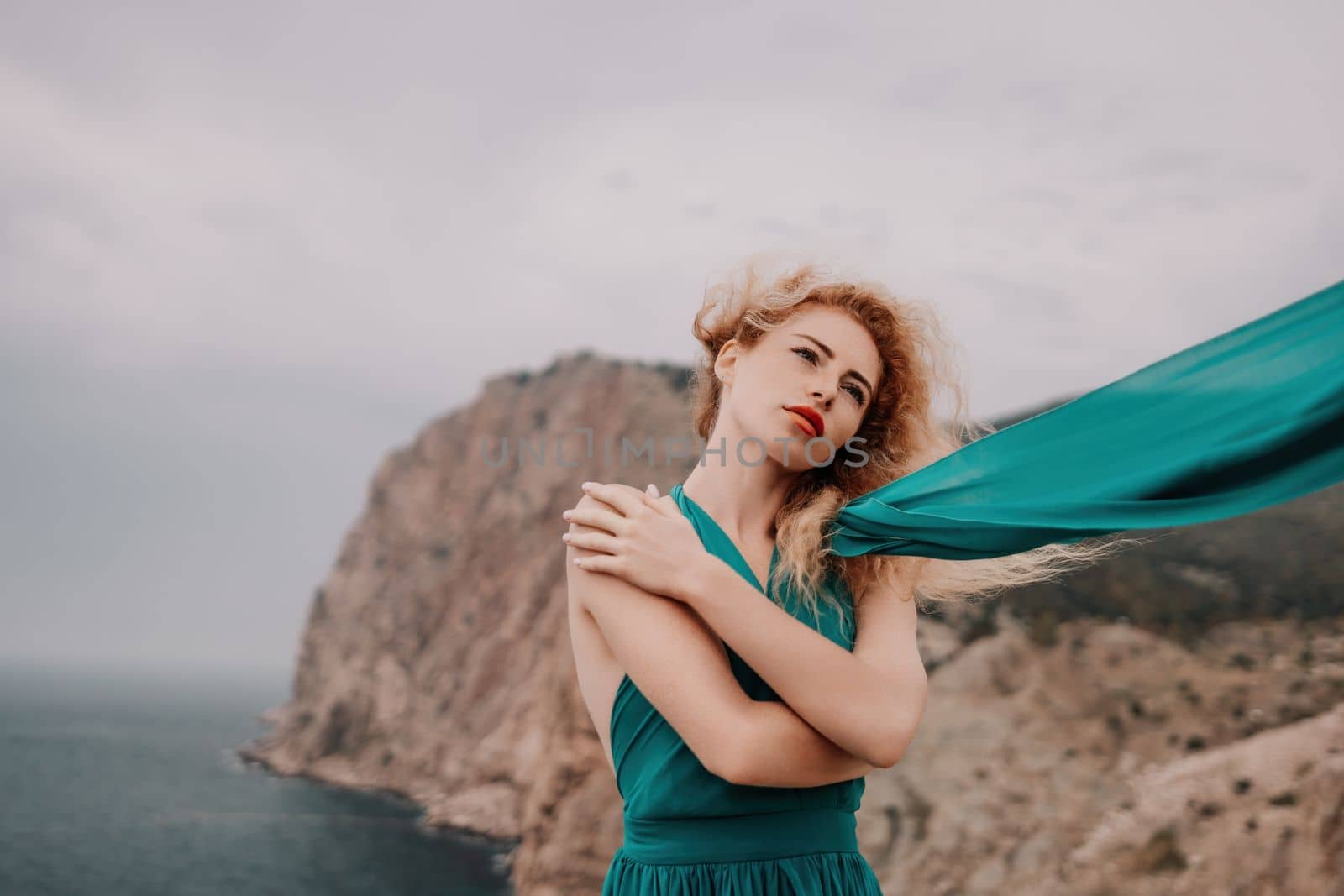 Redhead woman portrait. Curly redhead young caucasian woman with freckles looking at camera and smiling. Close up portrait cute woman in a mint long dress posing on a volcanic rock high above the sea by panophotograph