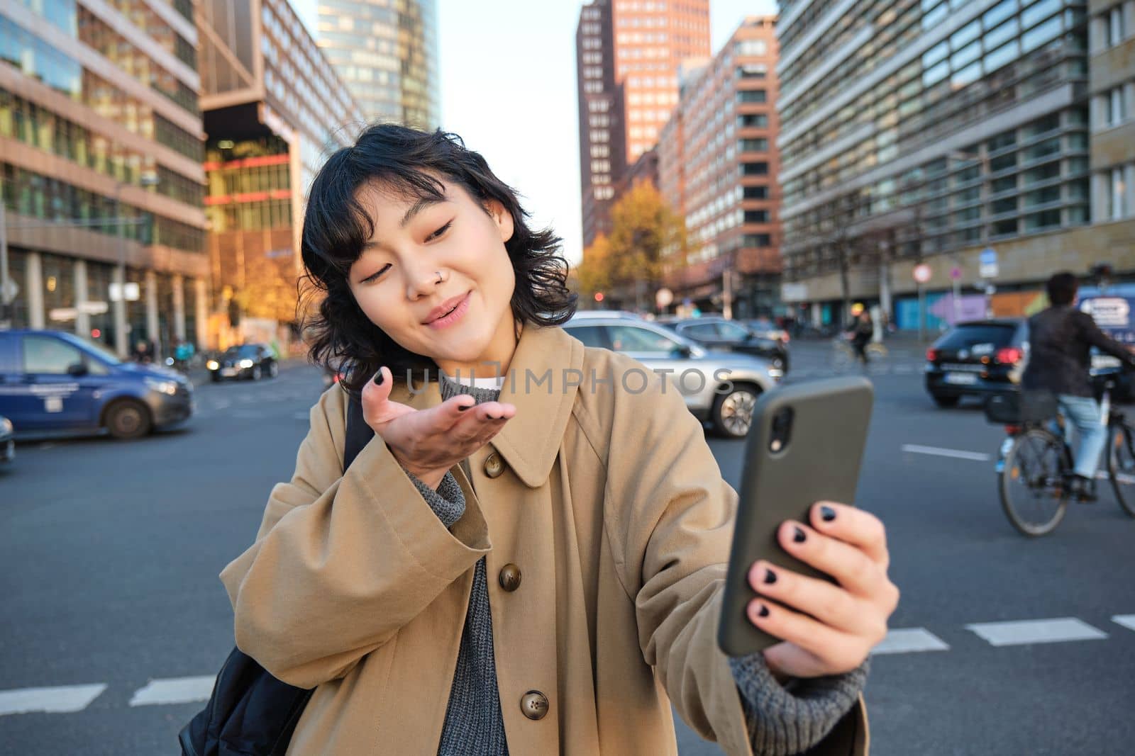 Cute korean girl, tourist takes selfie, video chats with friend and sends kisses to camera, records vlog on streets of city with smartphone.