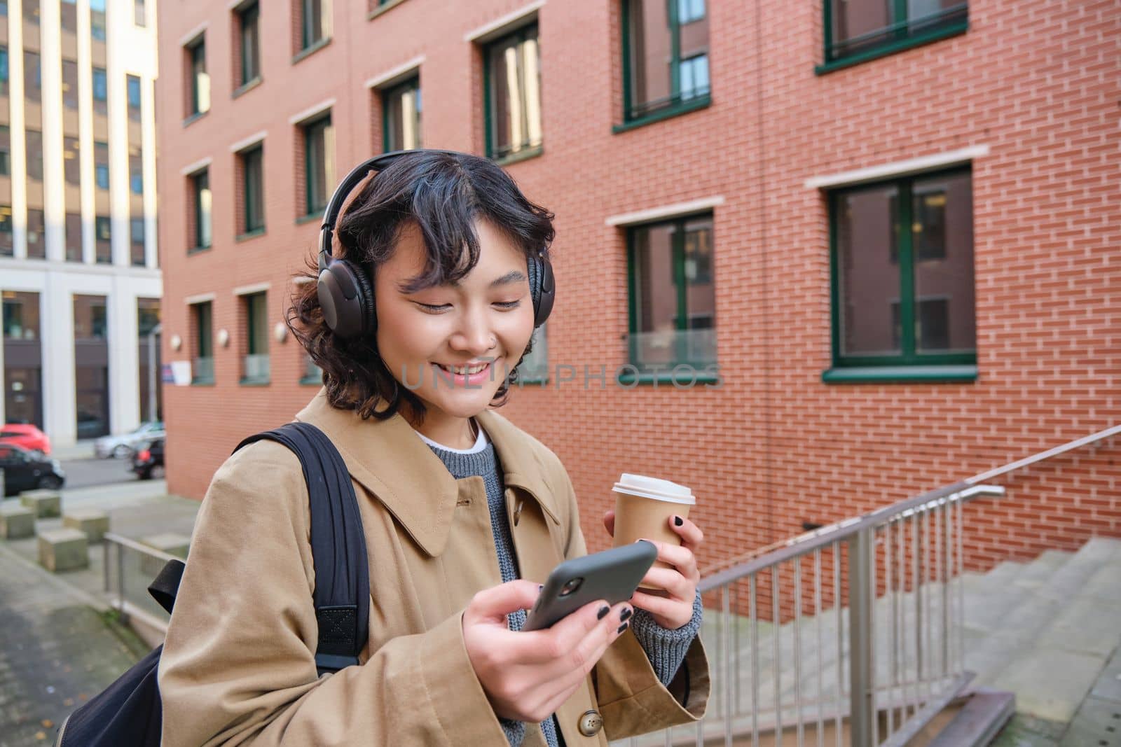 Young happy girl tourist, listens music in headphones, drinks takeaway and checks mobile phone, stands on street and smiles.