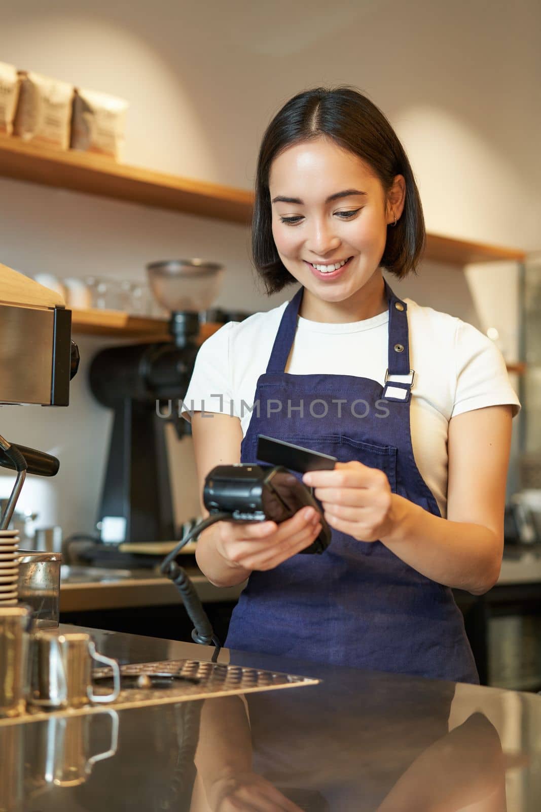 Portrait of smiling asian barista, coffee shop employee using POS terminal and credit card, helps client pay contactless in cafe.