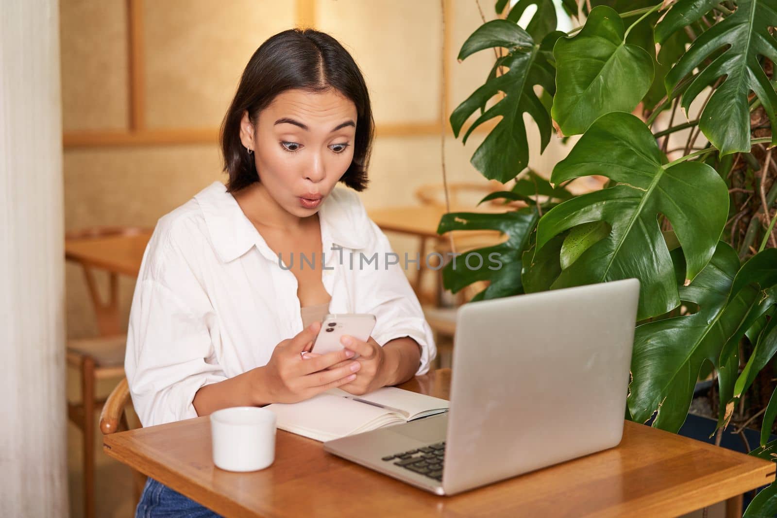 Woman in cafe sitting with laptop, looking surprised at smartphone screen, reading exciting good news.