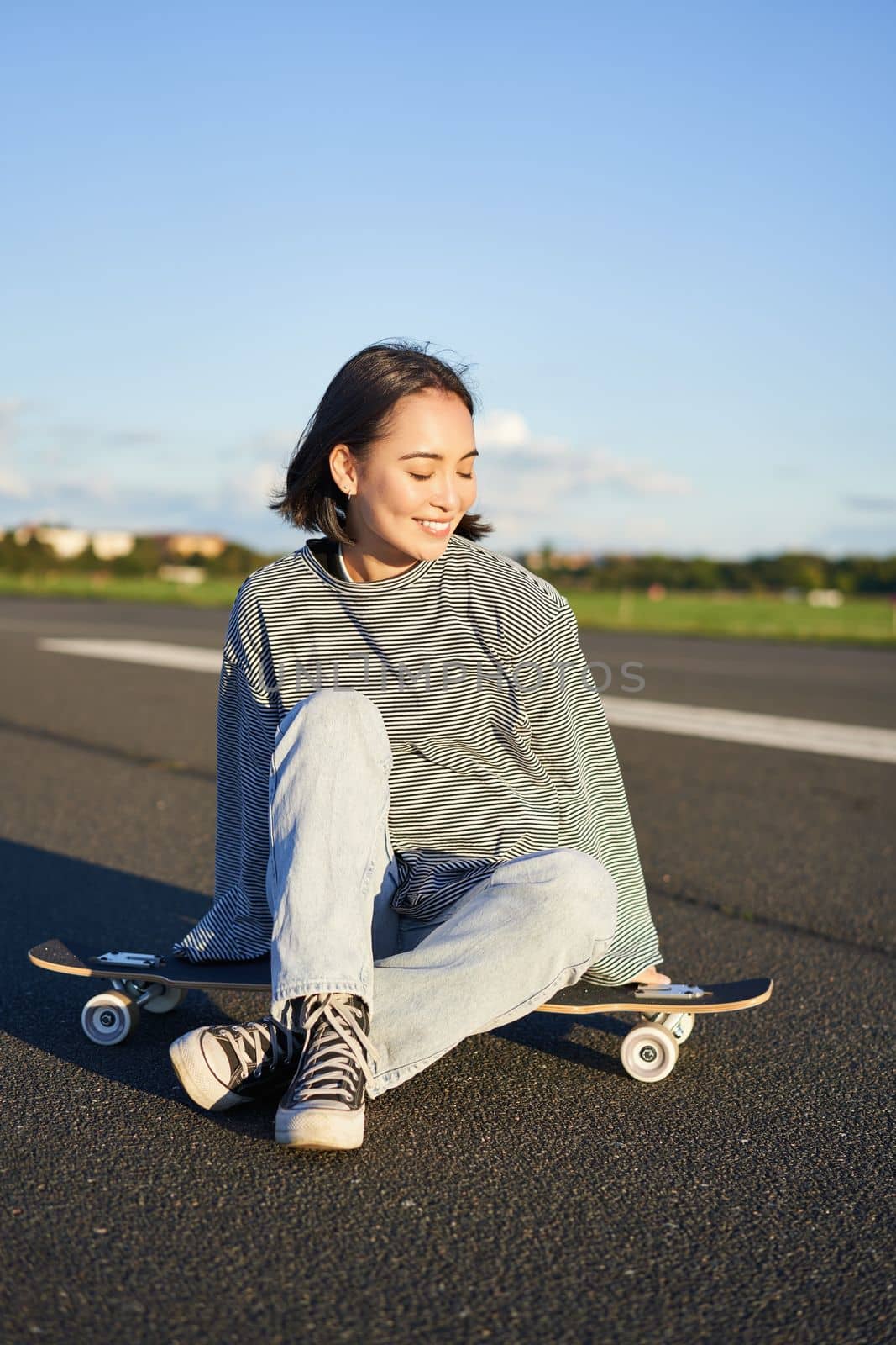 Cute smiling asian girl sits on longboard, skateboarding alone on an empty road. Copy space.