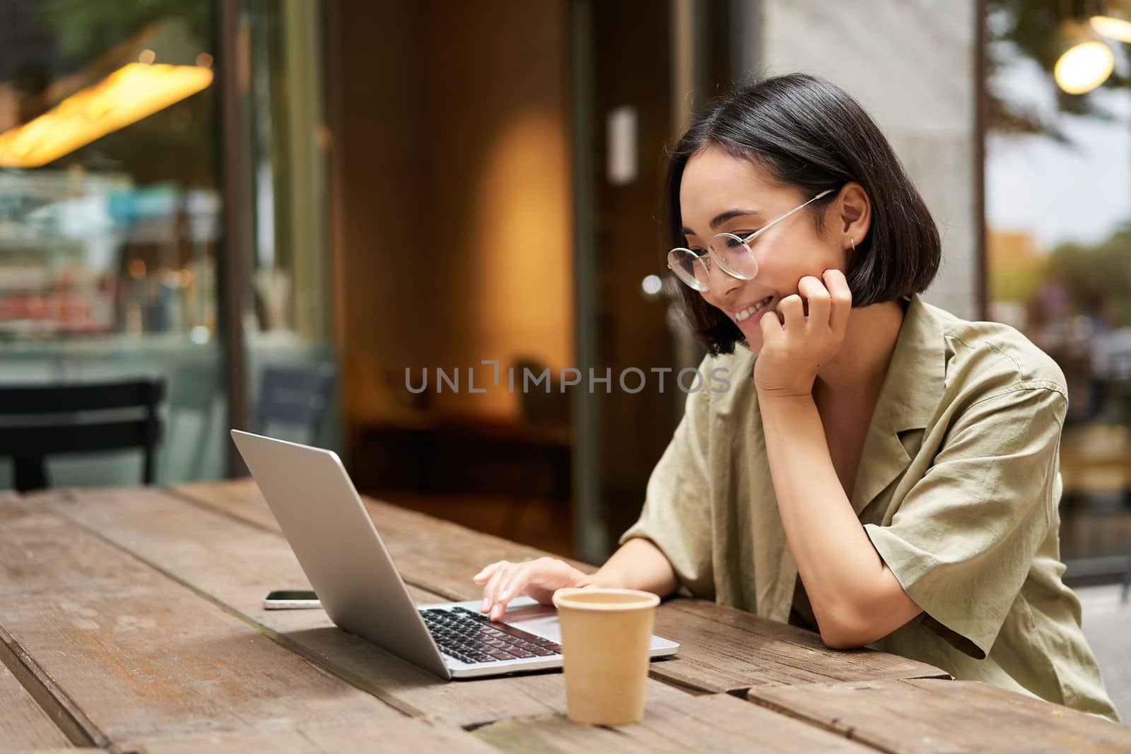 Portrait of smiling girl in glasses, sitting with laptop in outdoor cafe, drinking coffee and working remotely, studying online.