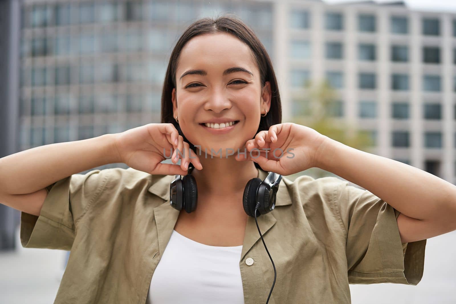 Portrait of smiling asian girl with headphones, posing in city centre, listening music.