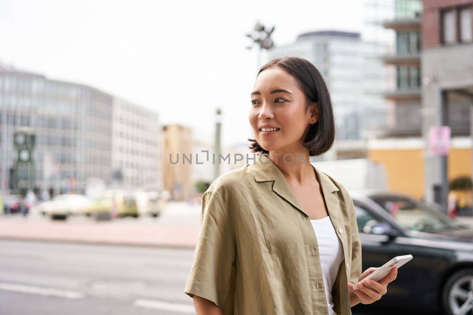 Young woman walking down the city street, turn behind, using map app navigator on smartphone, holding mobile phone.