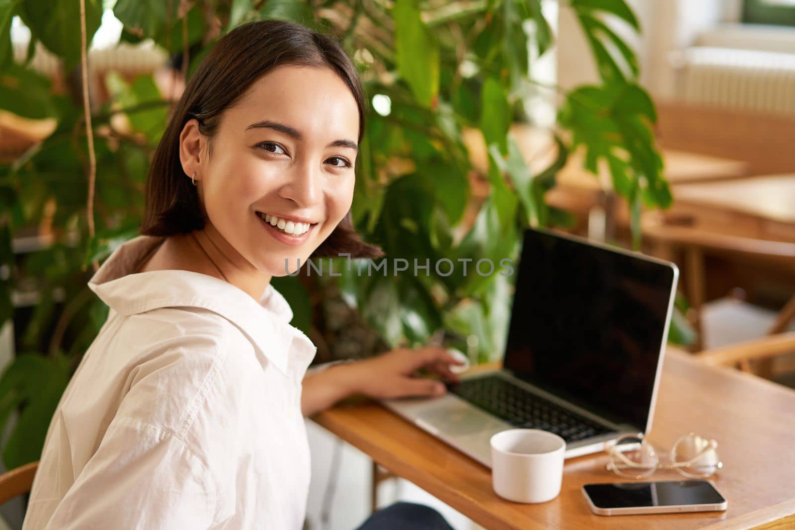 Young female entrepreneur, girl sits with laptop in cafe, works on remore from restaurant, drinks coffee and smiles.