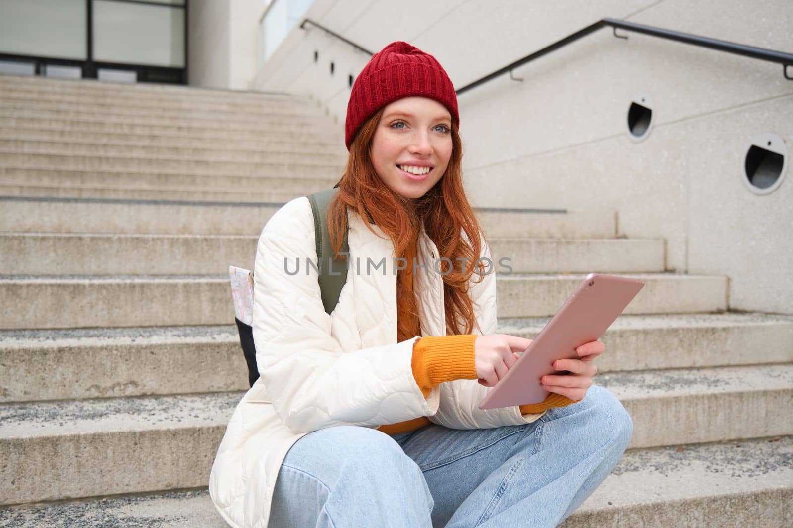 Young stylish girl, redhead female students sits on stairs outdoors with digital tablet, reads, uses social media app on gadget, plays games while waits on street.