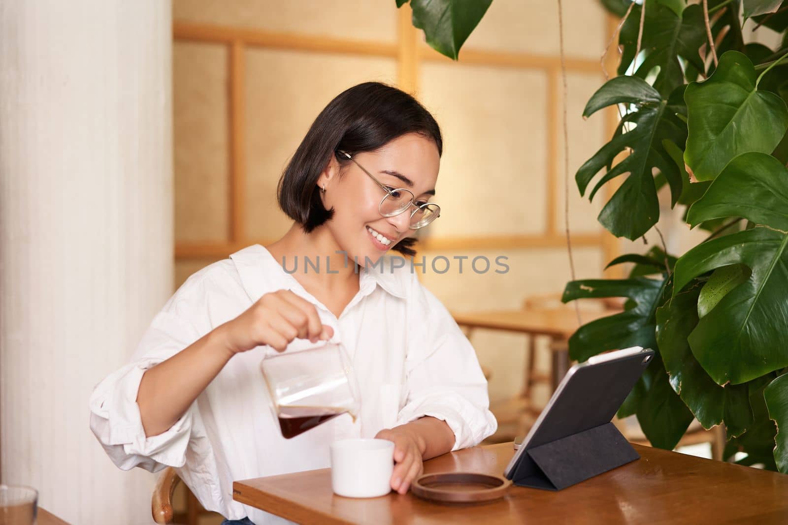 Freelance and remote workers. Smiling young woman pouring coffee in a cup, sitting in cafe and looking at digital tablet.
