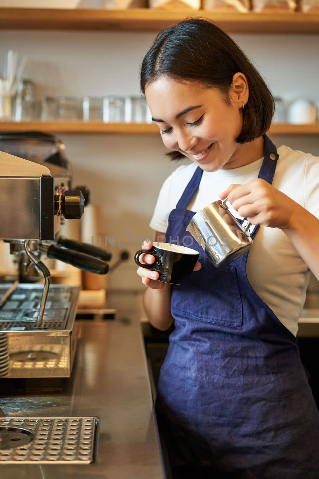 Close up of cute asian barista girl making cappuccino, doing latte art in cup with steamed milk, standing in coffee shop behind counter.