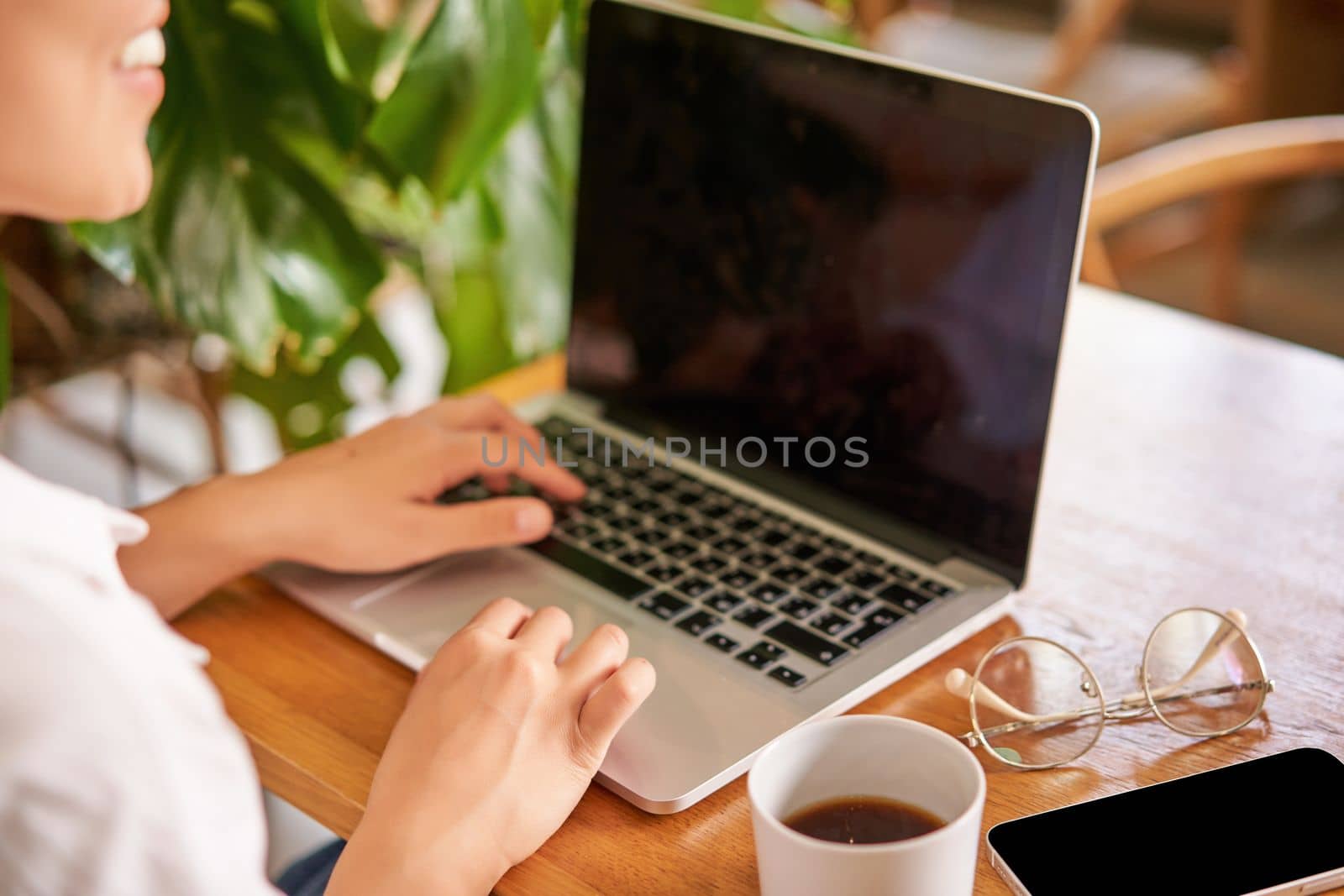 Cropped shot of laptop screen and female hands typing, cafe interior and cup of coffee with glasses on table.