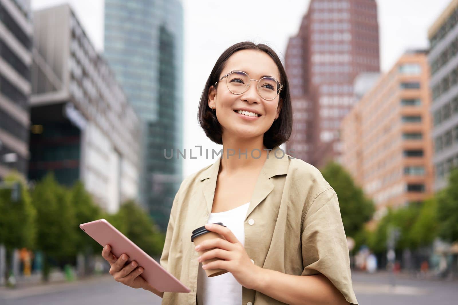 Portrait of happy young woman in glasses, standing on street with cup of coffee and tablet by Benzoix