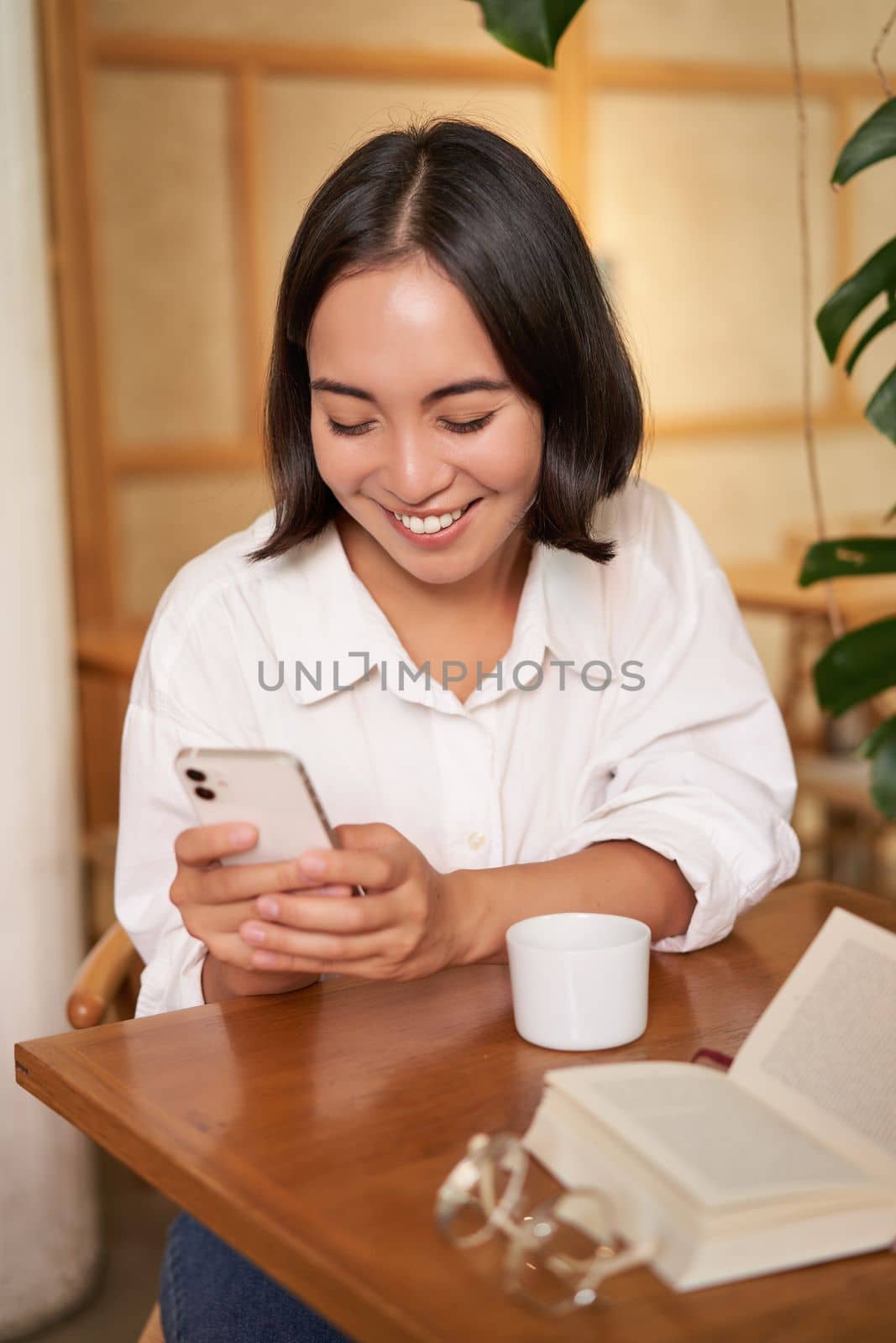 Portrait of modern girl sits with coffee in cafe, smiling while looking at smartphone, reading book in restaurant and using mobile phone app.