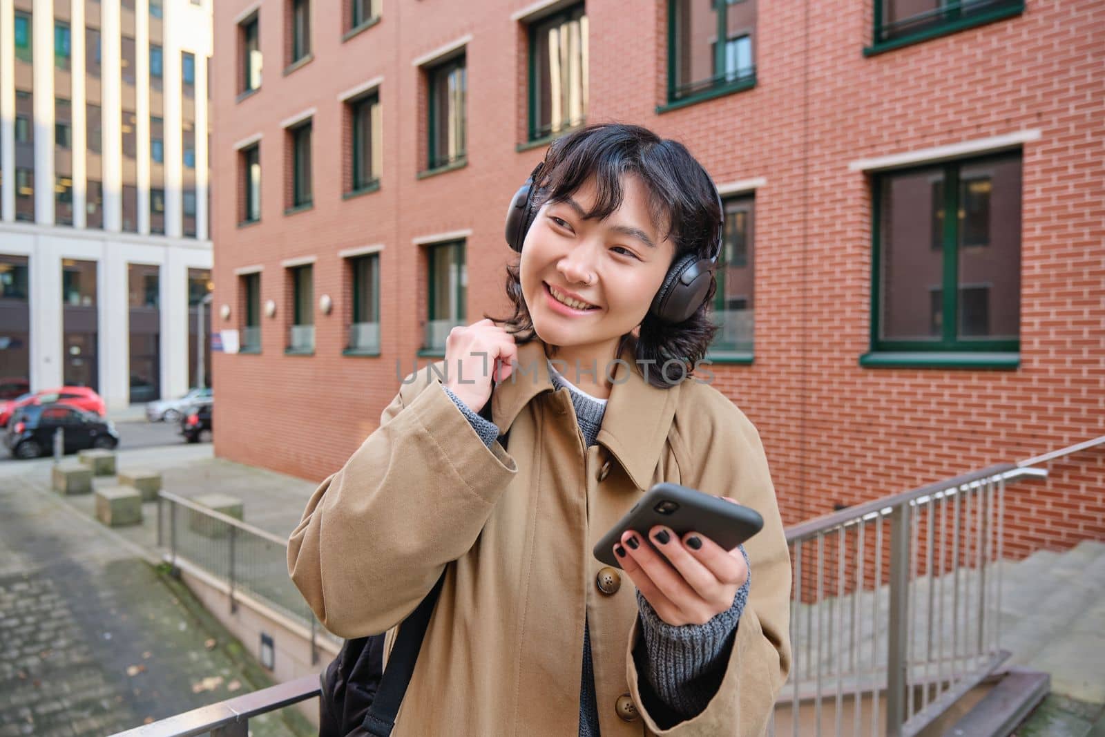 Young happy woman in trench coat, listens music in headphones, holds smartphone, uses mobile application to find route in new city, stands on street with backpack.