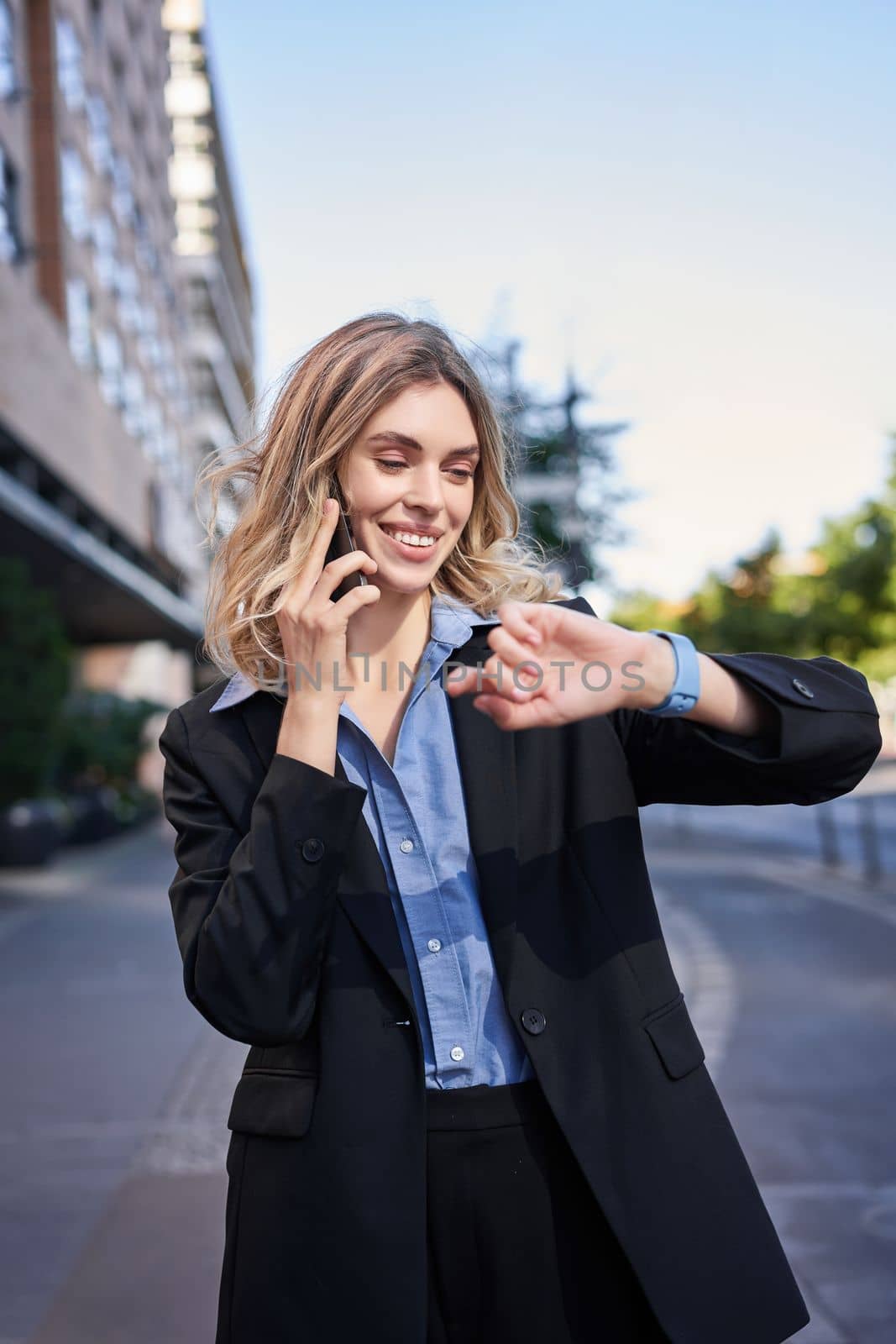 Portrait of confident businesswoman going on a meeting, standing on street, talking on mobile phone and checking time on digital watch.