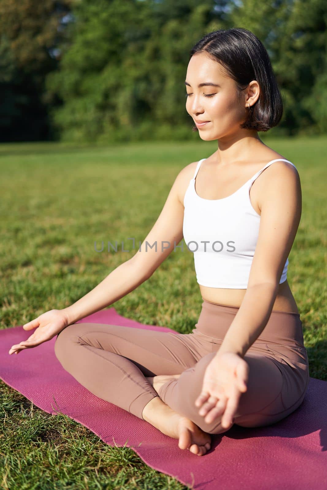 Woman meditating in park on rubber mat, sitting on green lawn and practice yoga, concept of sport and wellbeing.