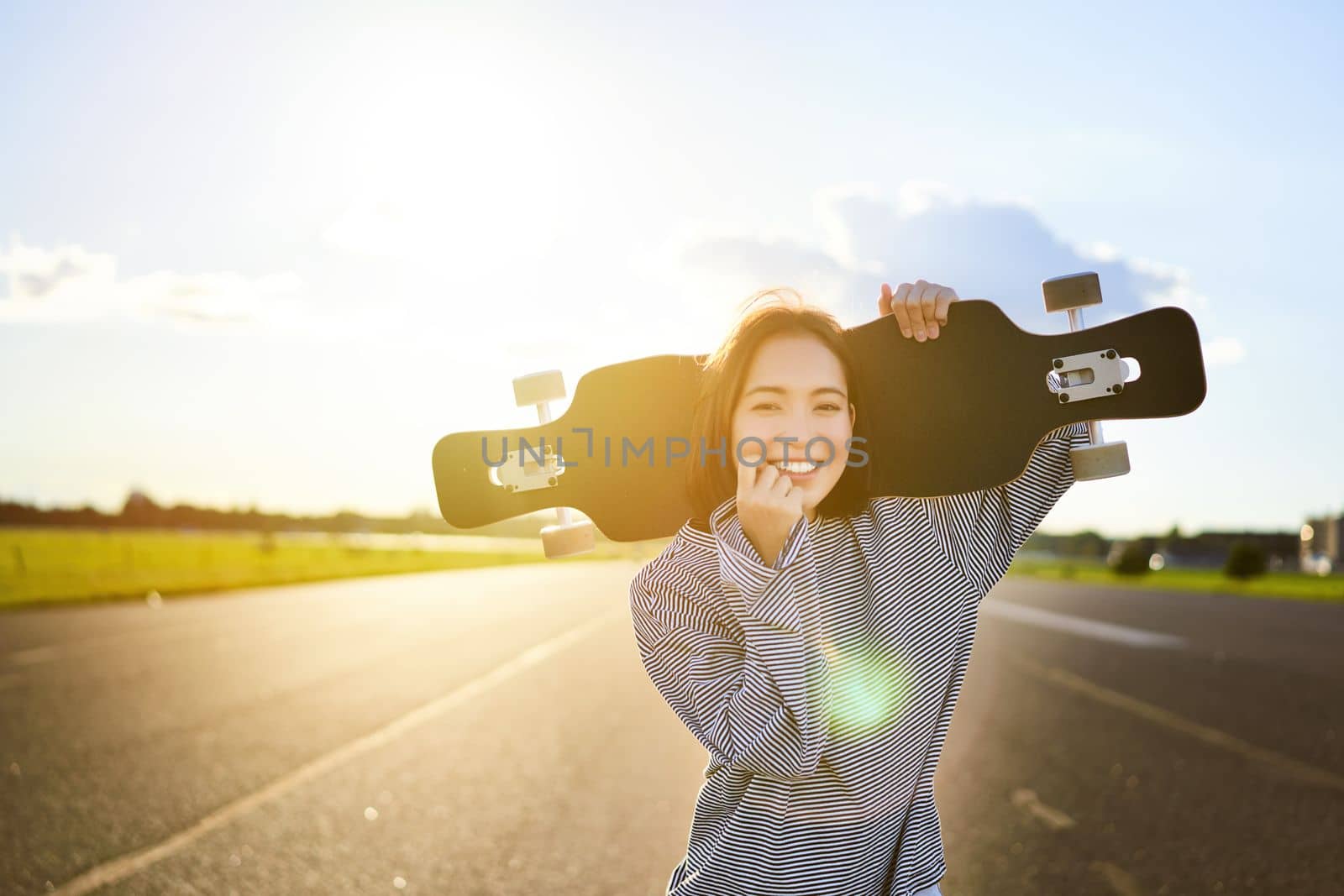 Happy skater girl walking towards the camera, sunbeams shining at lense. Young woman skating on cruiser, holding longboard on shoulders.
