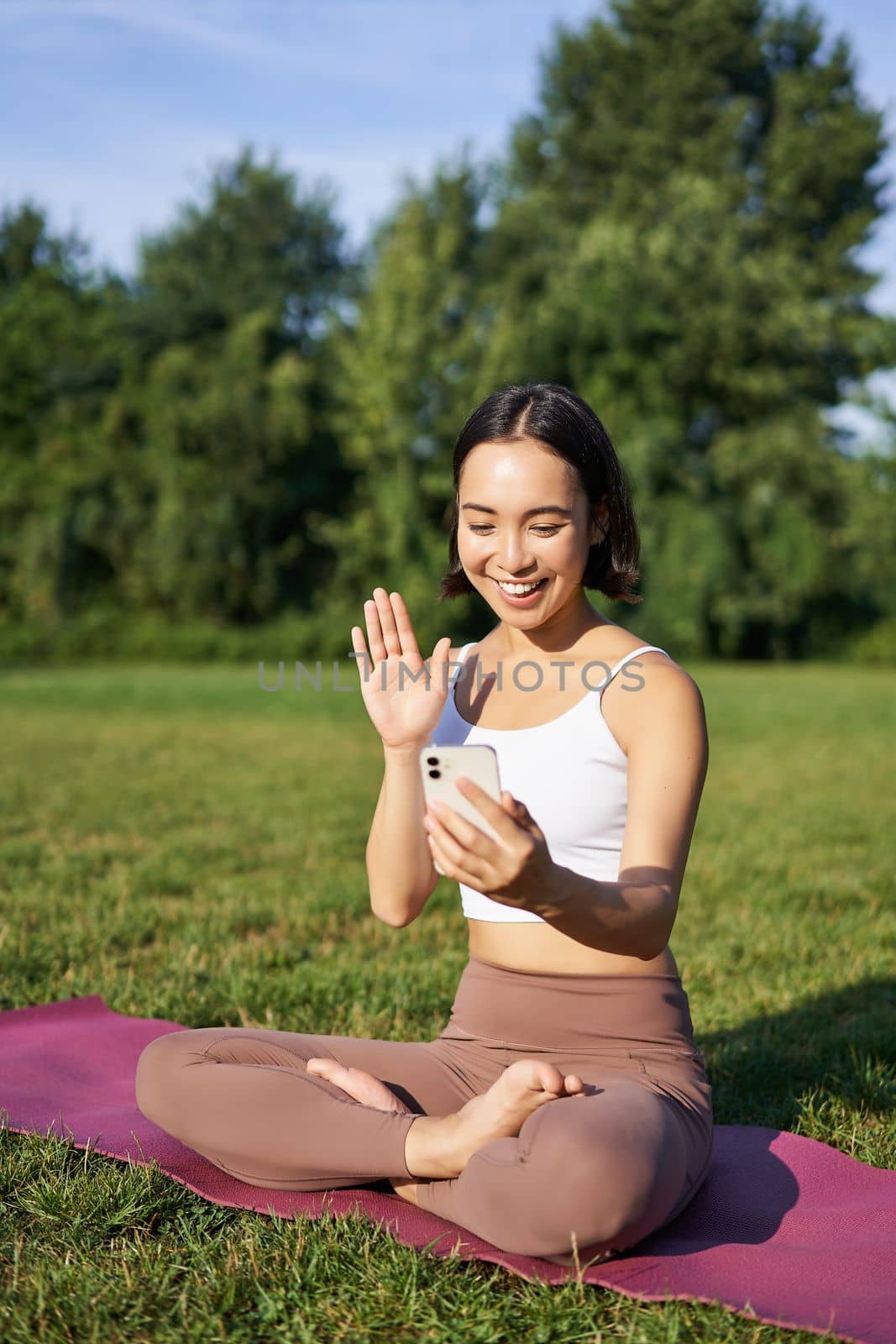 Portrait of asian girl sitting on mat in park, talking to smartphone, meditating online with yoga instructor on fresh air by Benzoix