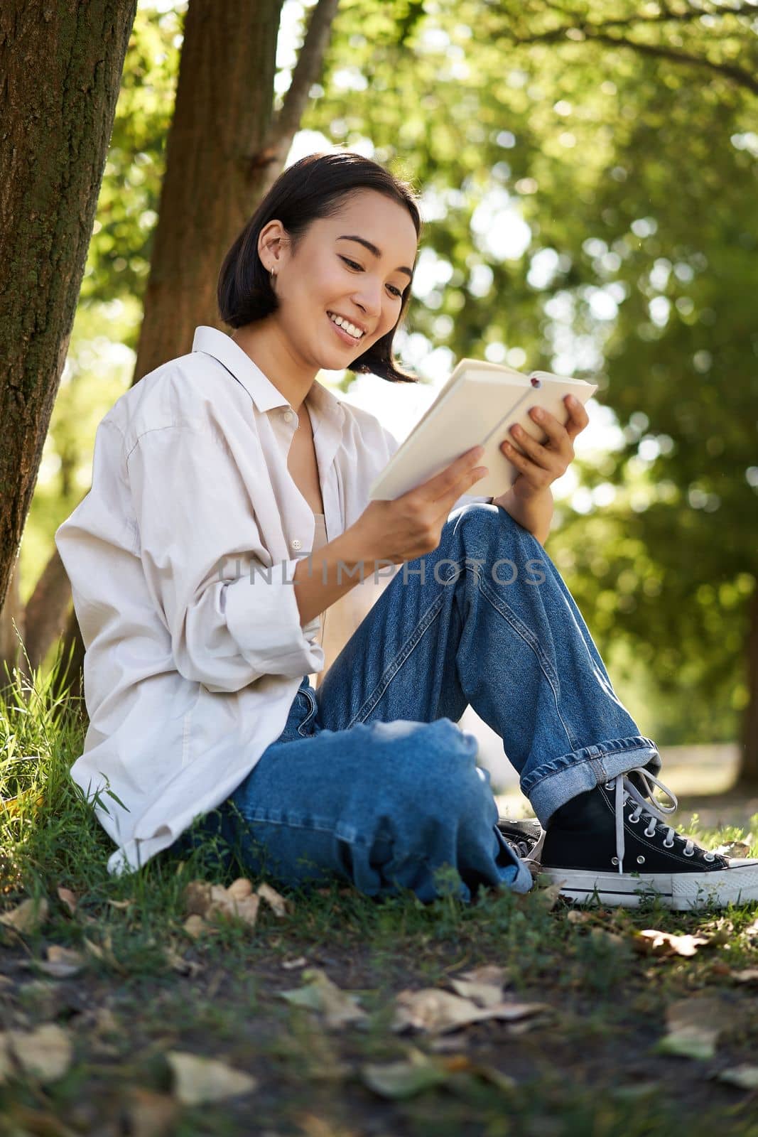 Beautiful young asian girl, student sits in park under tree and reading book, smiling, enjoying warm summer day outdoors by Benzoix