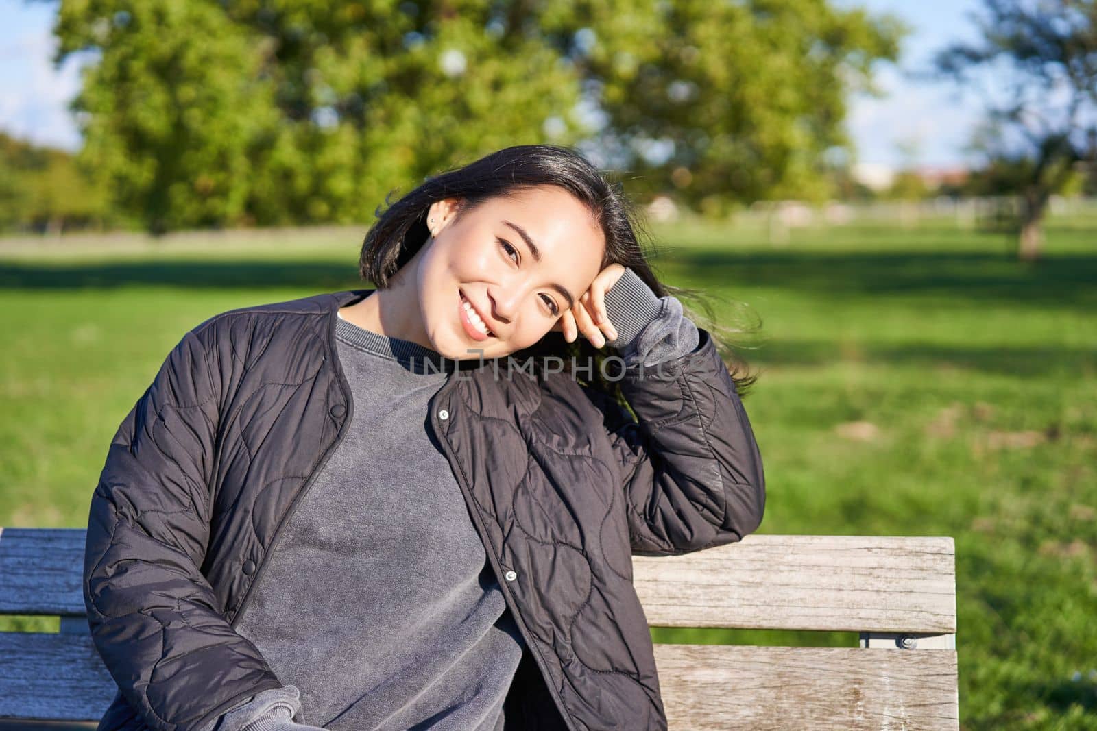 Beauty portrait of smiling asian girl with dark short hair, sitting on bench in park and gazing at camera.
