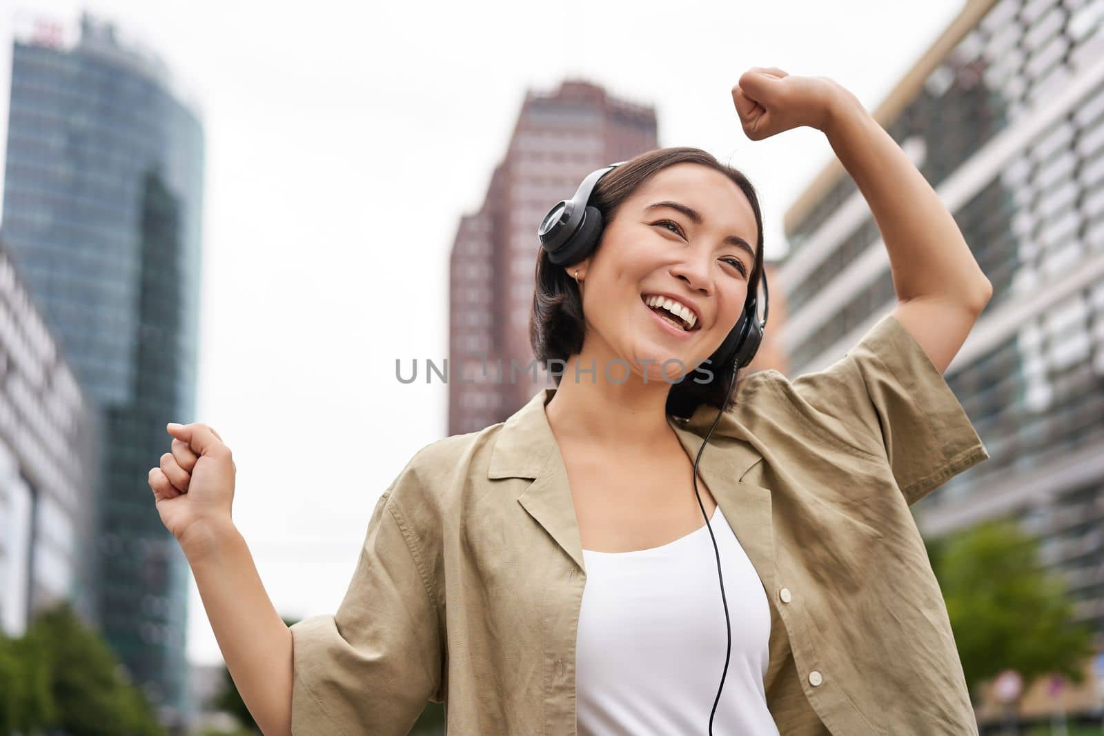 Dancing asian girl listening music in headphones, feeling happy in city, enjoying outdoors.