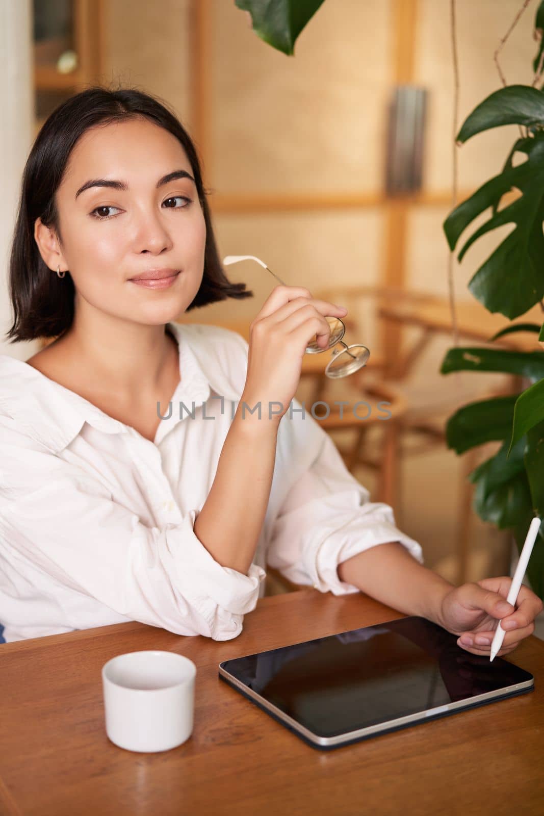 Smiling young female creator, creative girl drawing on digital tablet with graphic pen, drinking coffee in cafe by Benzoix