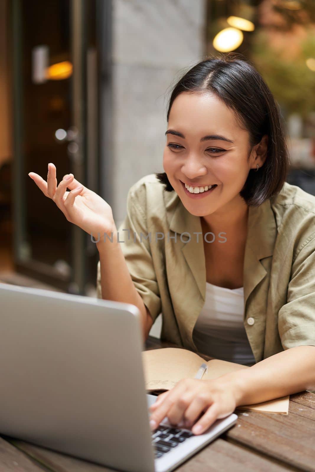 Vertical shot of young brunette girl chatting on video chat, having online meeting, talking to laptop, sitting in cafe.