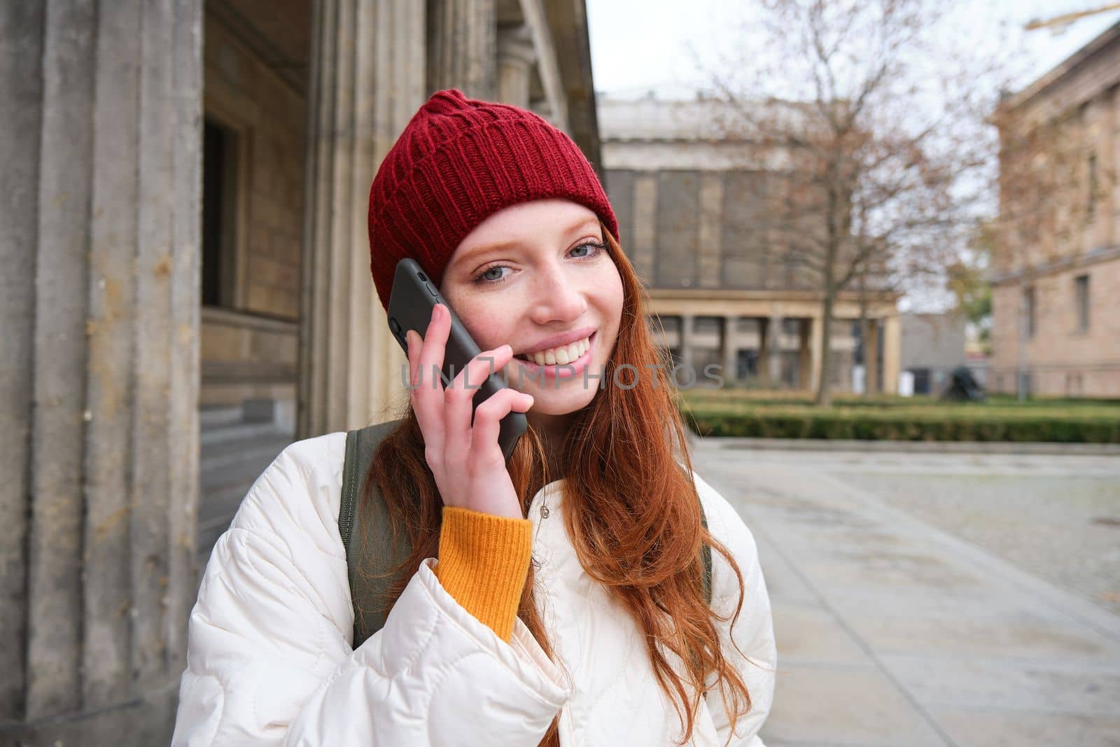 Smiling young redhead woman listens to voice message, makes a phone call, walks on street and talks to someone on smartphone.