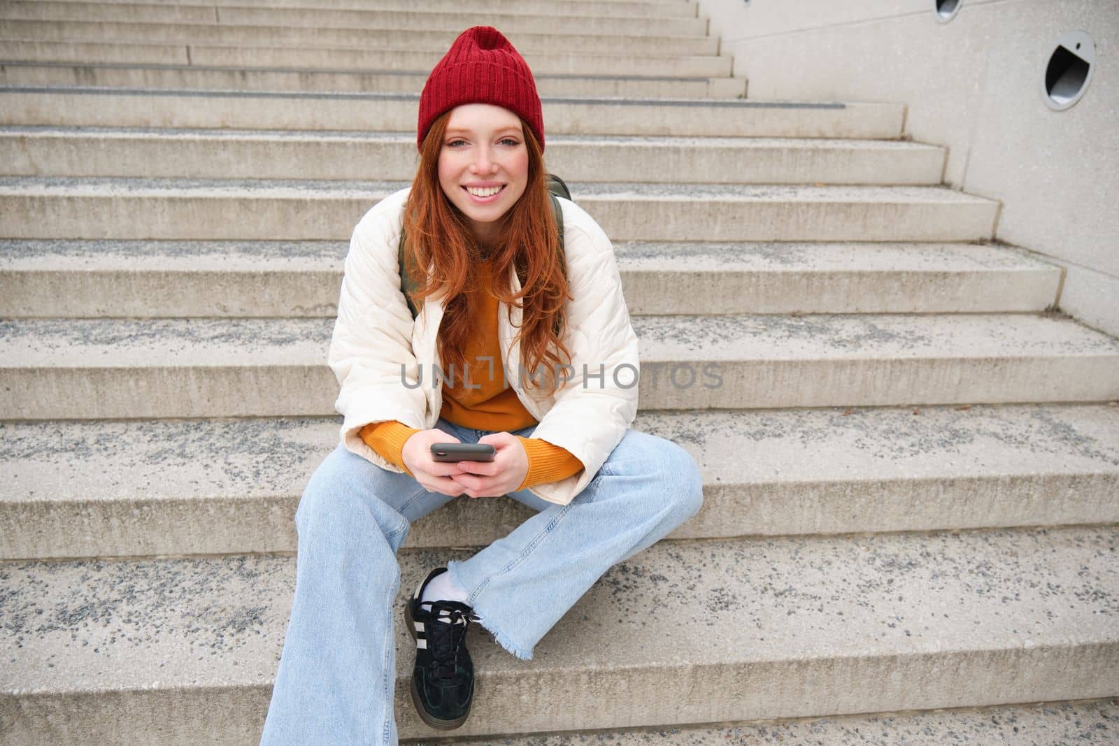 Hipster ginger girl, redhead woman sits on stairs with smartphone, waits for someone and messages on social media on mobile phone app. People and technology concept