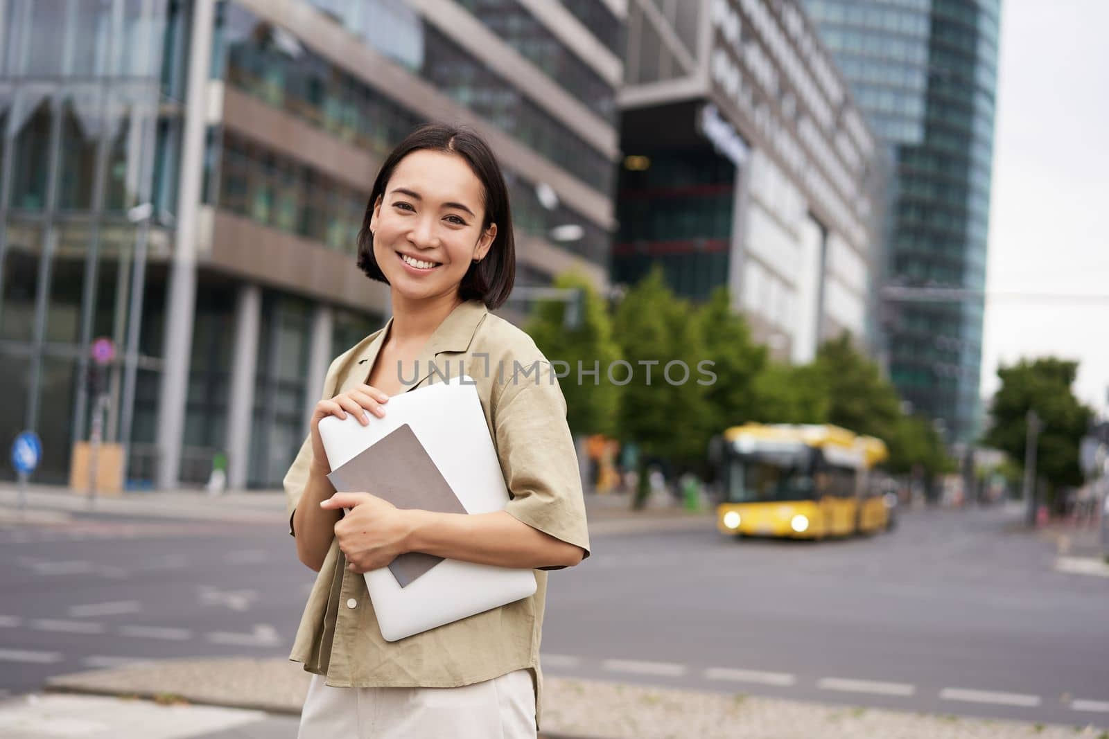 Beautiful asian girl smiles as commutes to work, stands on street with laptop and notebook.