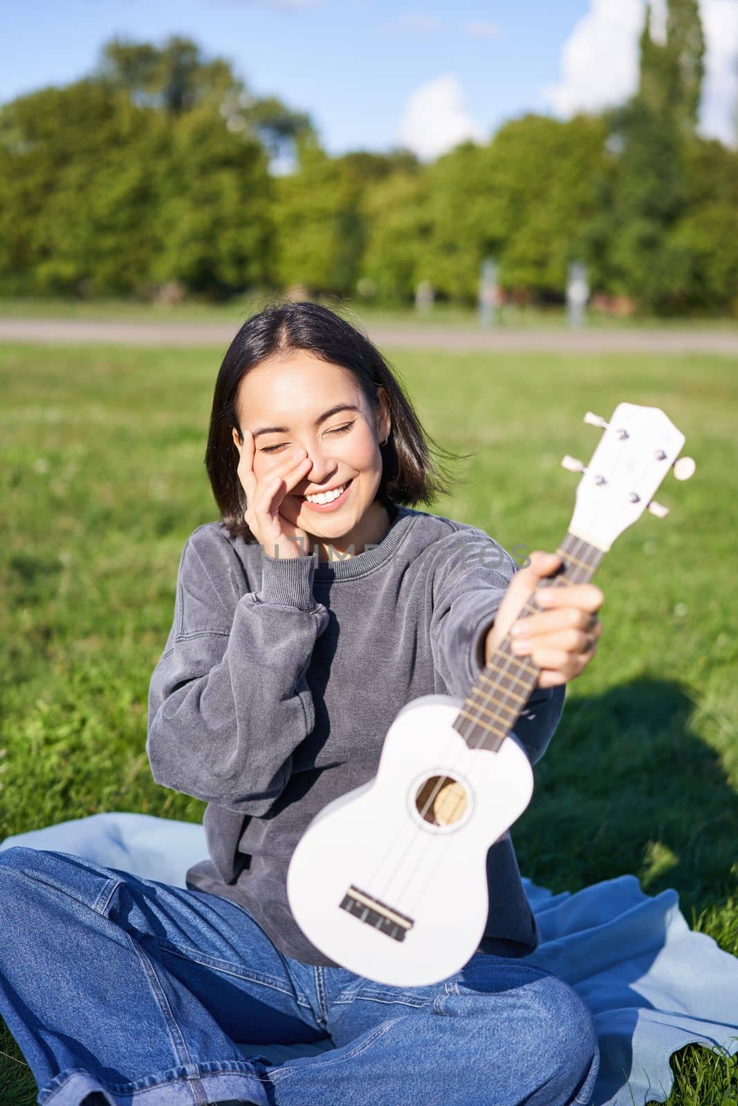 Music and instruments. Smiling asian girl shows her white ukulele, sits in park and plays small guitar by Benzoix