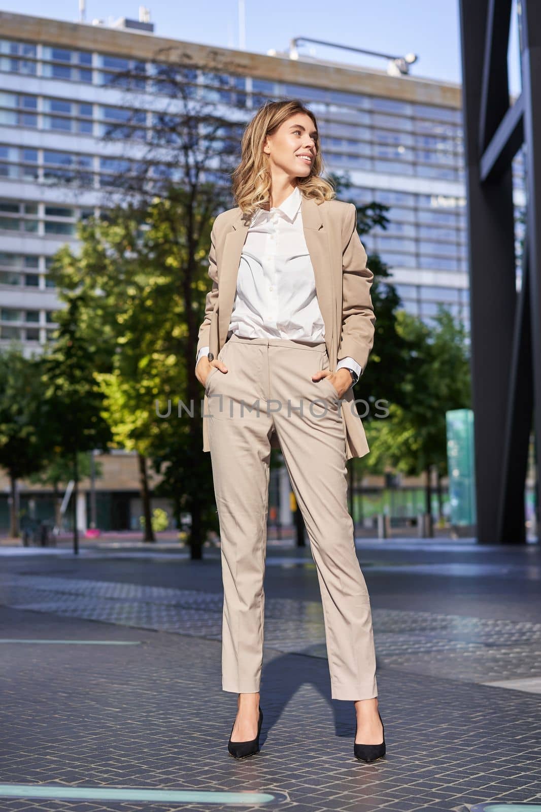 Vertical portrait of young confident businesswoman in high heels and beige suit, looking aside, smiling, holding hands in pockets.