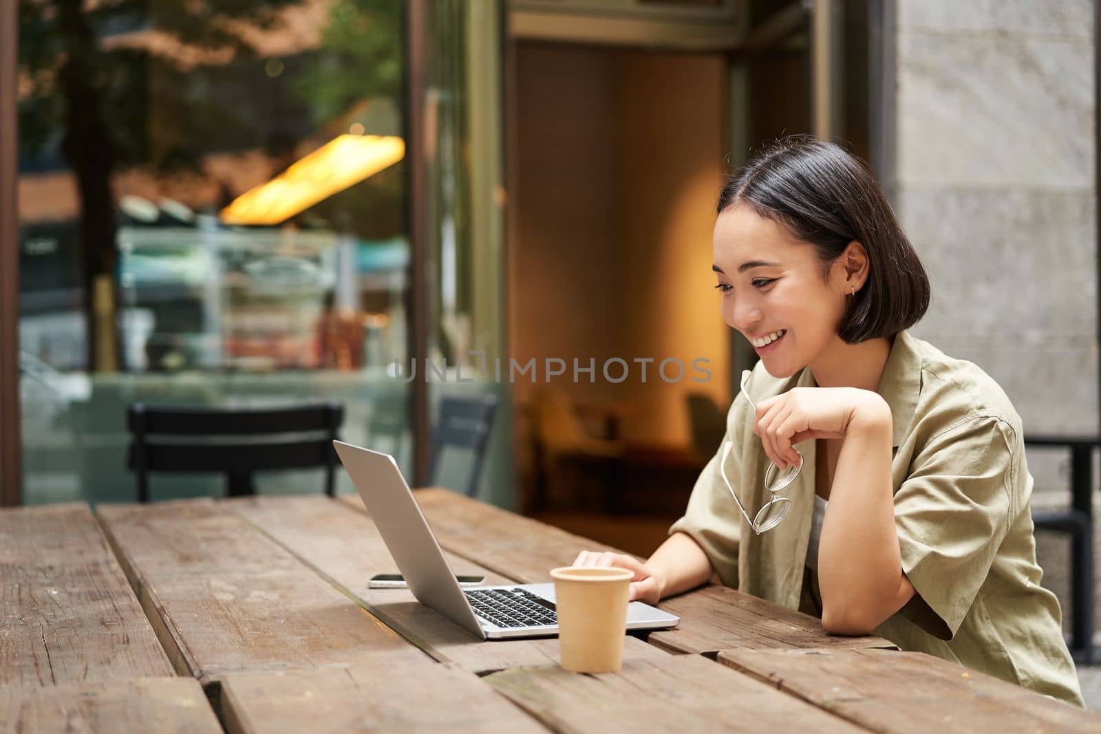 Portrait of asian woman looking at laptop, video chat, talking with someone via computer camera, sitting in cafe and drinking coffee, online meeting.