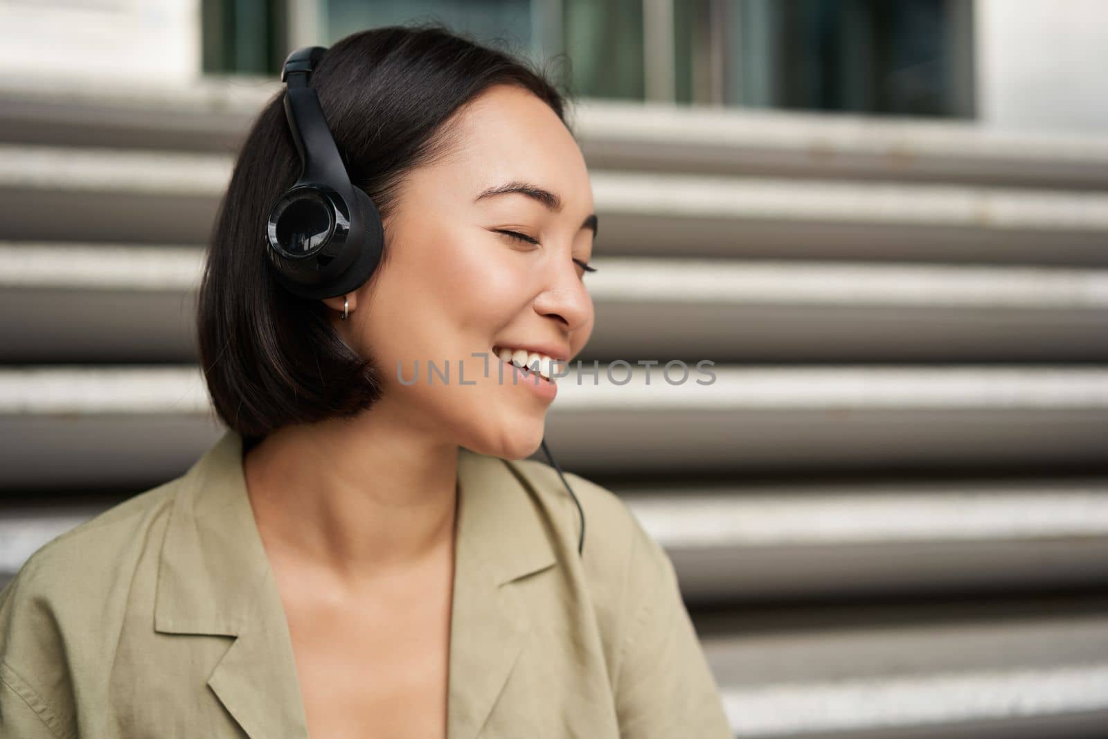Young asian woman listening to music in headphones, sitting outside on street, smiling.