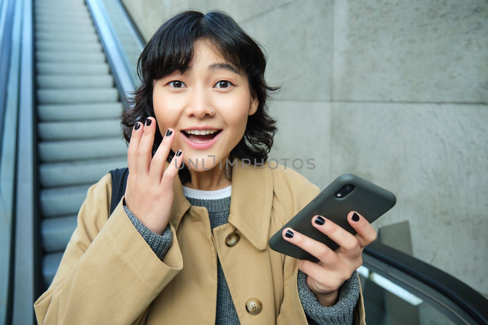 Joyful and positive korean girl, celebrates, looks surprised, goes down escalator with smartphone and looks amazed by smth.