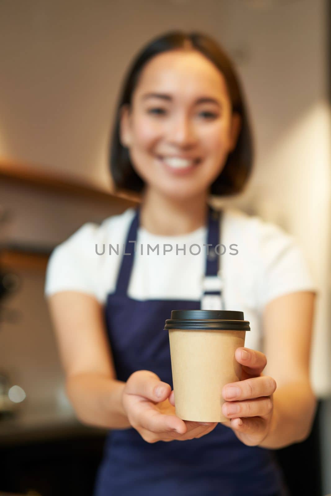 Smiling asian barista girl, giving takeaway coffee cup, prepare takeout order to guest in cafe, wearing apron uniform.