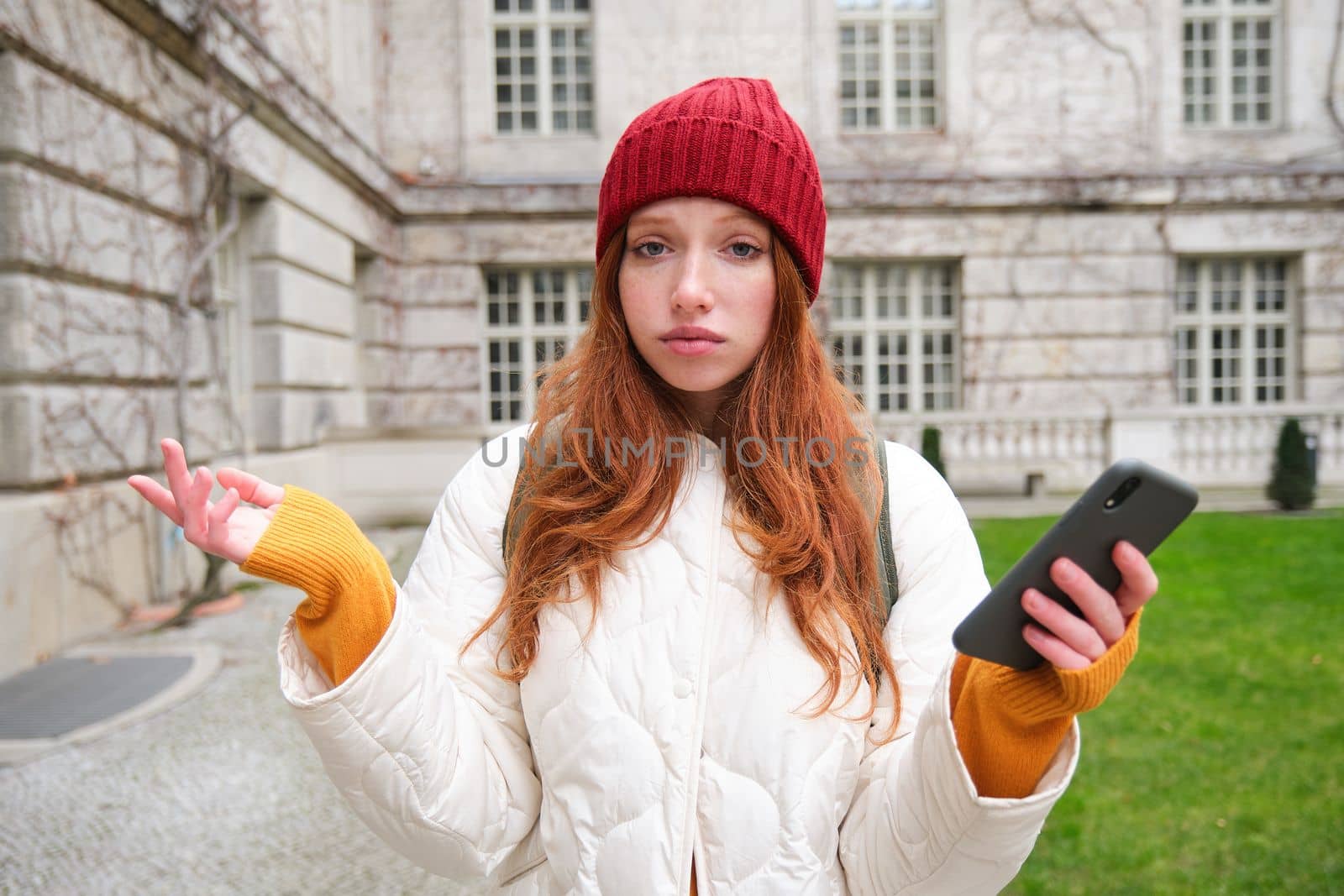 Portrait of confused redhead girl with smartphone, shrugs shoulders with puzzled, clueless face expression, tourist got lost in city.