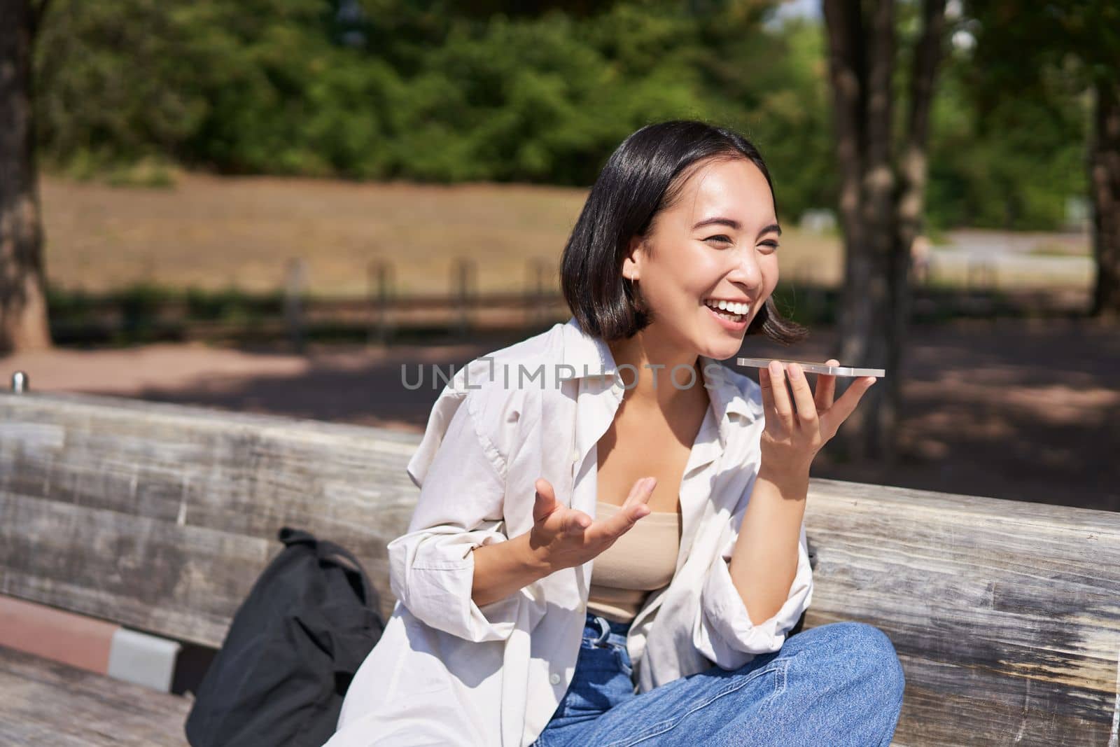 Portrait of happy asian girl records voice message, talking via speakerphone on smartphone, sitting relaxed on bench in park on sunny day.