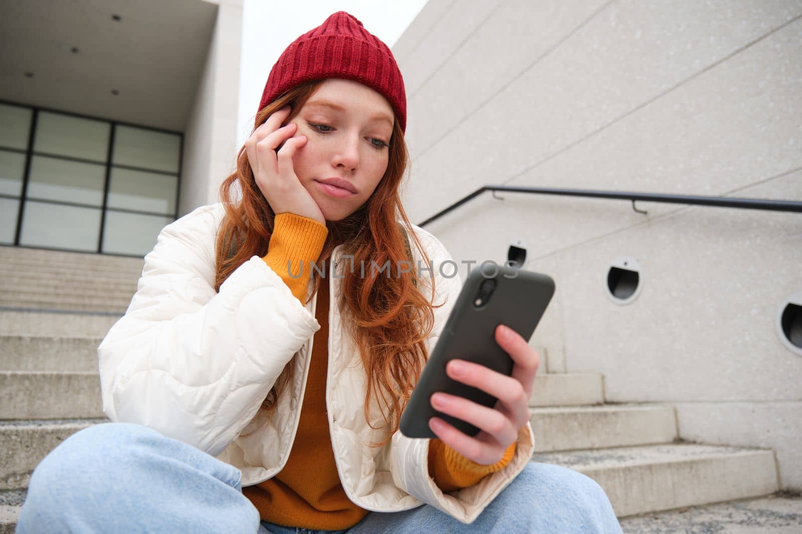 Mobile phones and people. Young stylish redhead girl sits on stairs with telephone, uses smartphone app, reads smth online.