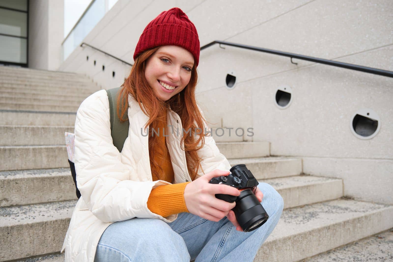 Smiling redhead girl photographer, checks her shots, holds camera and looks at screen, takes photos outdoors, walks around street and does streetstyle shooting by Benzoix