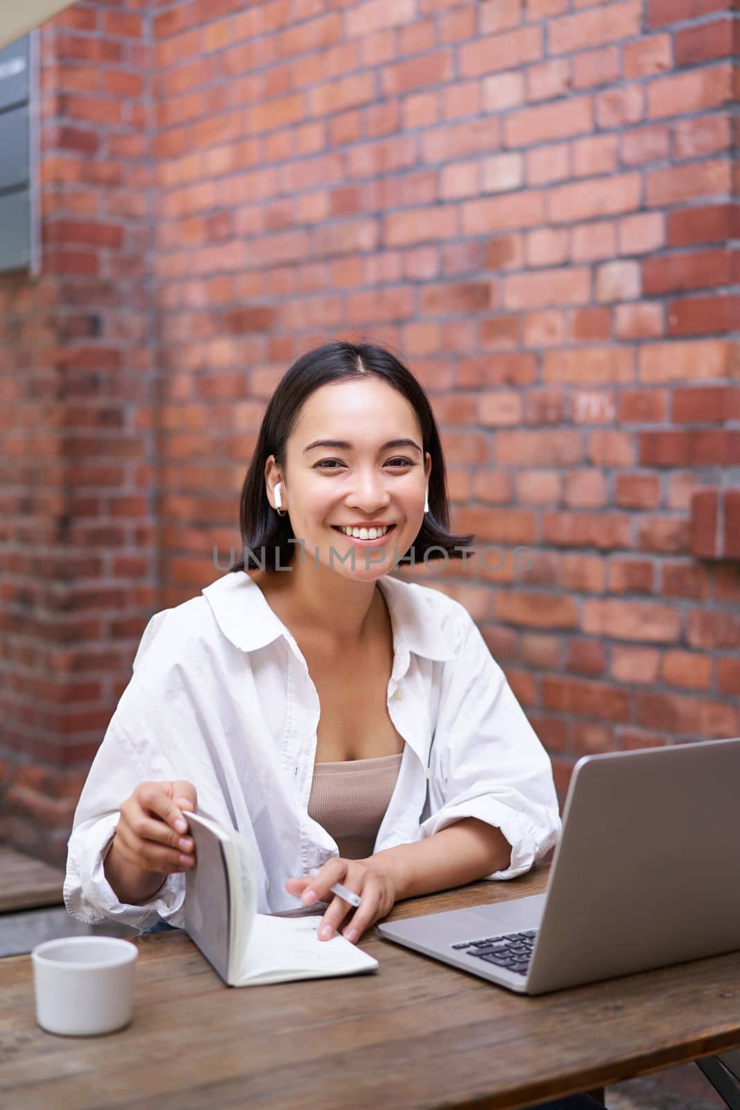 Stylish asian girl works in coworking with laptop, makes notes, uses wireless earphones and smiles, sits near brick wall in office.
