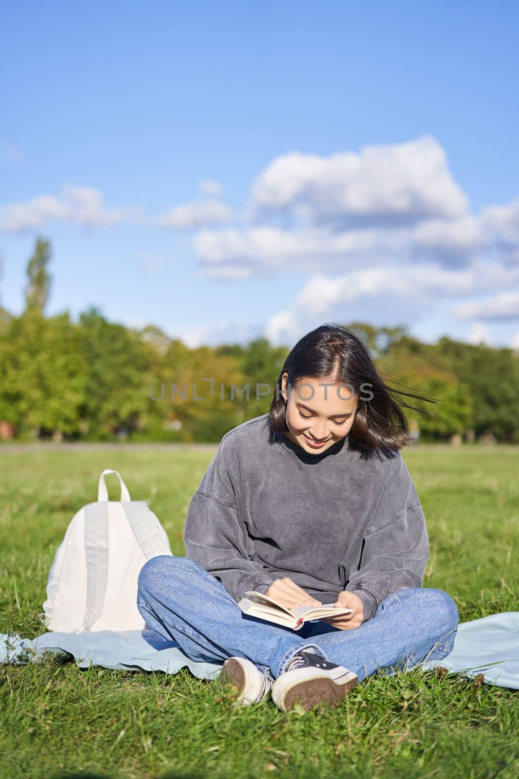 Vertical shot of cute teen girl sits in park on grass with backpack and her book, reading alone outdoors.