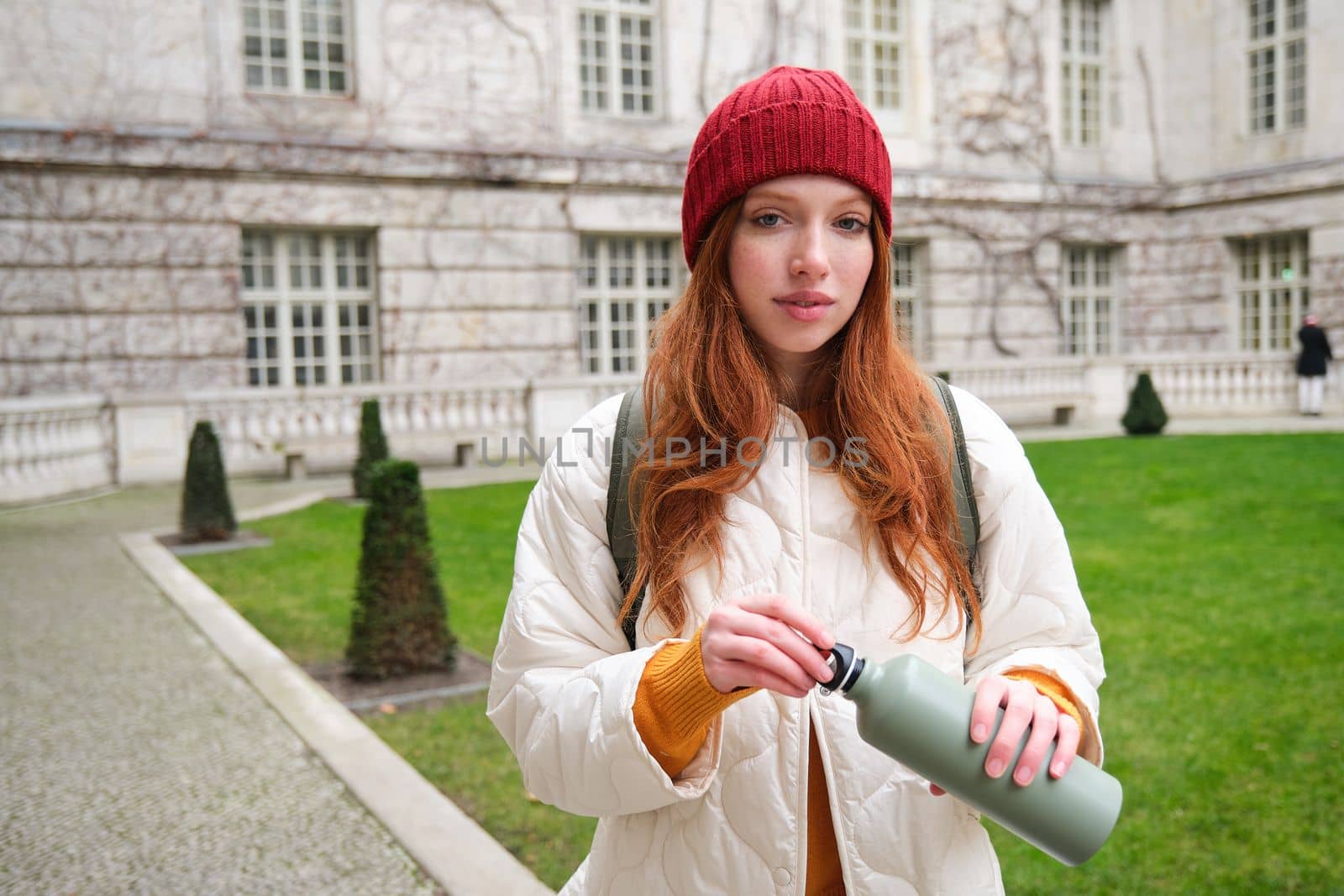 Portrait of happy young woman, tourist with backpack sightseeing, drinking hot tea from thermos, holding flask and smiling by Benzoix